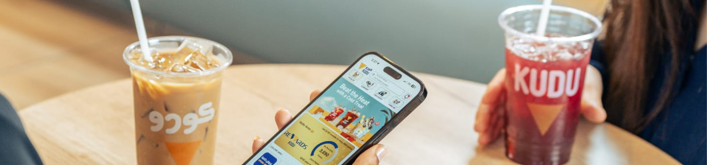 Two people sit at a wooden table, enjoying drinks from KUDU. One woman scrolls through KUDU's rewards app.